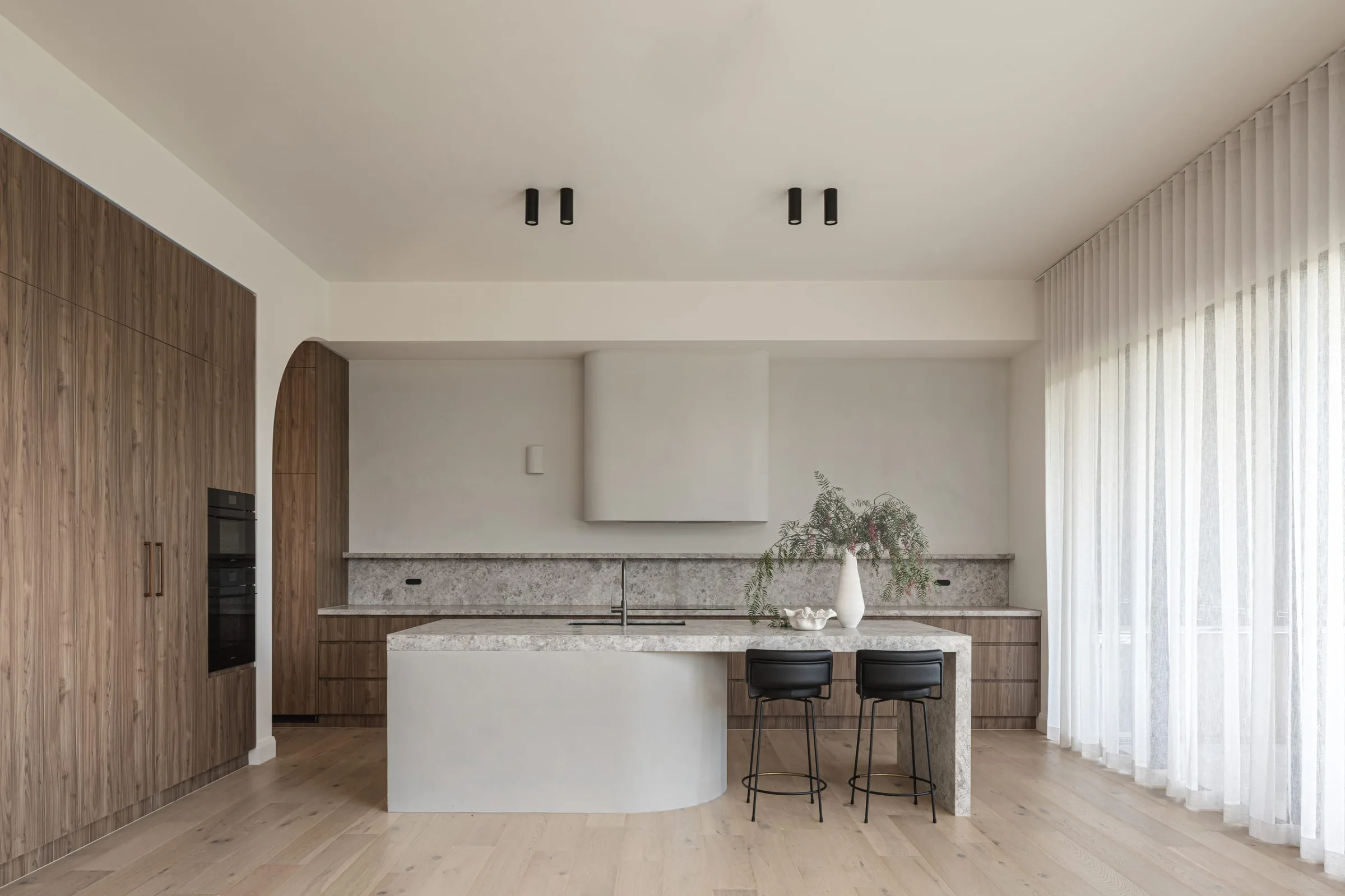 Kitchen with a curved white island, timber joinery and sheer curtains, Whites Hill House